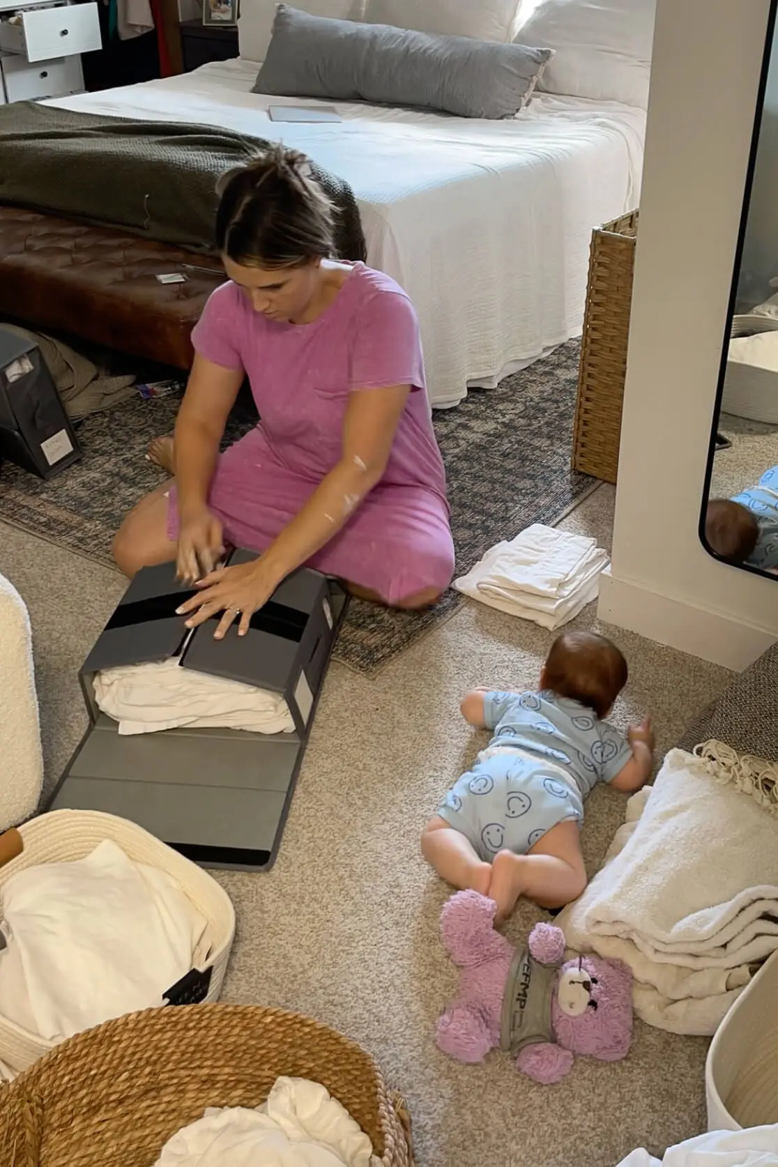 Woman folding and organizing bed linens into a storage bin while her baby plays nearby on the carpet during a home organization project.